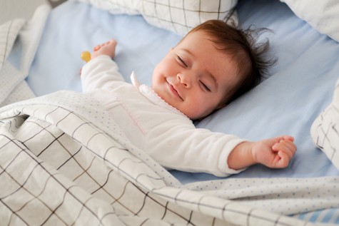 Baby smiling in bed with eyes closed and arms out.